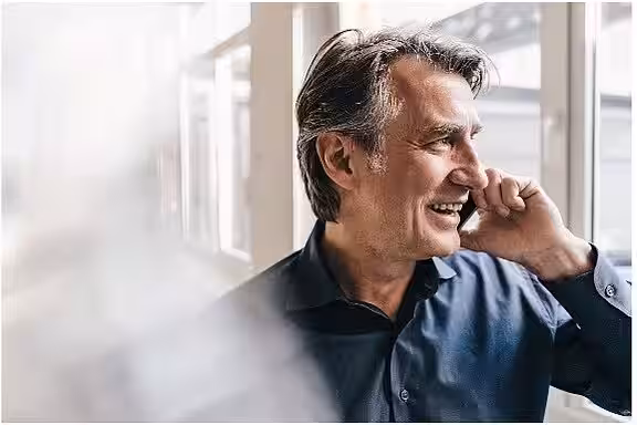 Auf dem Bild ist ein erwachsener Mann zu sehen der aus dem Fenster schaut und ein Telefon am Ohr hat. Er führt ein Gespräch mit einem LIFTON-Berater und lacht dabei.